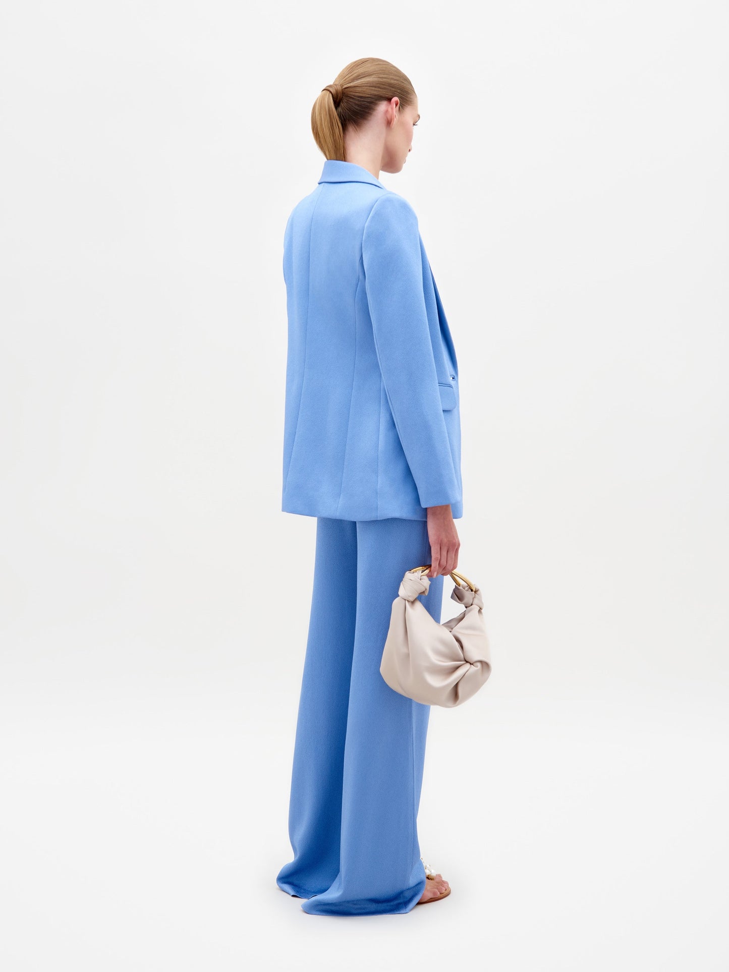 A woman in a light blue Rebeca Jacket with hammered satin, single-button closure, and matching knotted top and pants holds sunglasses against a plain white background.