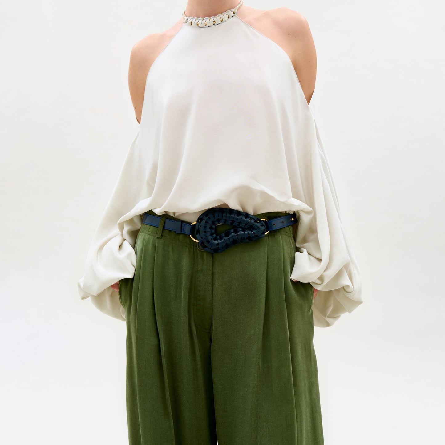 Person wearing a white blouse and green pants with a decorative belt on a plain background