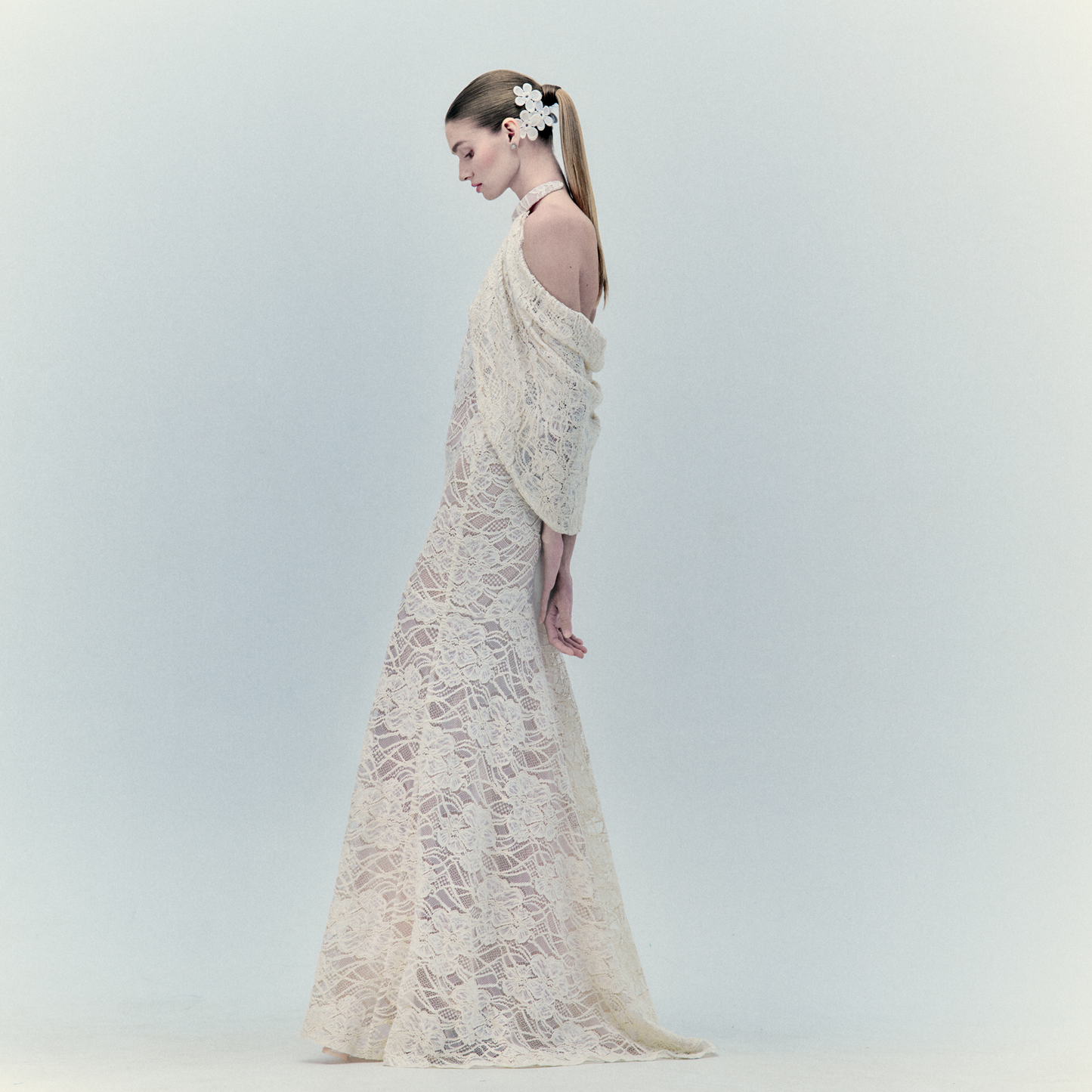 A woman stands in profile wearing the Rita Dress, a long lace maxi with a sheer pattern and halter neckline. Her hair is tied back with white flowers, while the minimal light background highlights the dress’s ethereal charm.