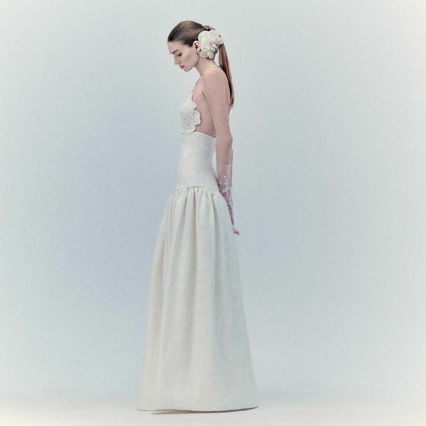 A woman in profile wears the elegant Ruth Dress—a long white maxi—against a pale background. Her hair is styled in a ponytail with a large floral appliqué as she looks down, hands at her sides.