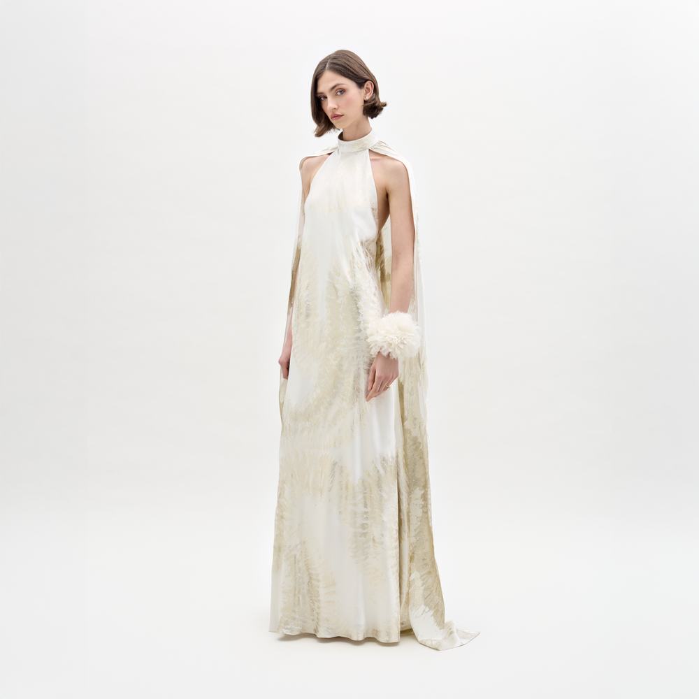 A woman poses in the Sabina Dress, a white and beige flowing gown featuring a high neckline and cape-like sleeves, set against a plain backdrop.