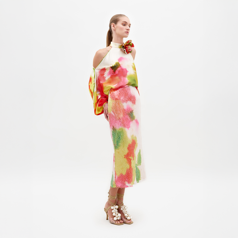 A person stands facing slightly to the side, wearing a colorful off-shoulder top and the Sarla Skirt Spring Blooms midi skirt, paired with high heels adorned with large pearl decorations; pre-order now to ensure it ships by February 15th, 2025.