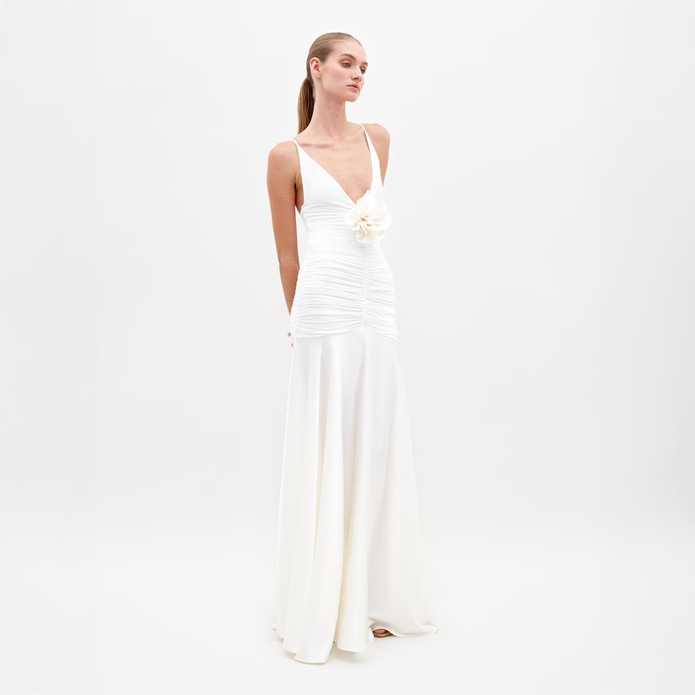 A woman in the Sana Dress White, featuring a sleeveless design with a deep V-neck and floral detail at the waist, stands against a plain white background. Pre-order now to ensure delivery by February 15th, 2025.