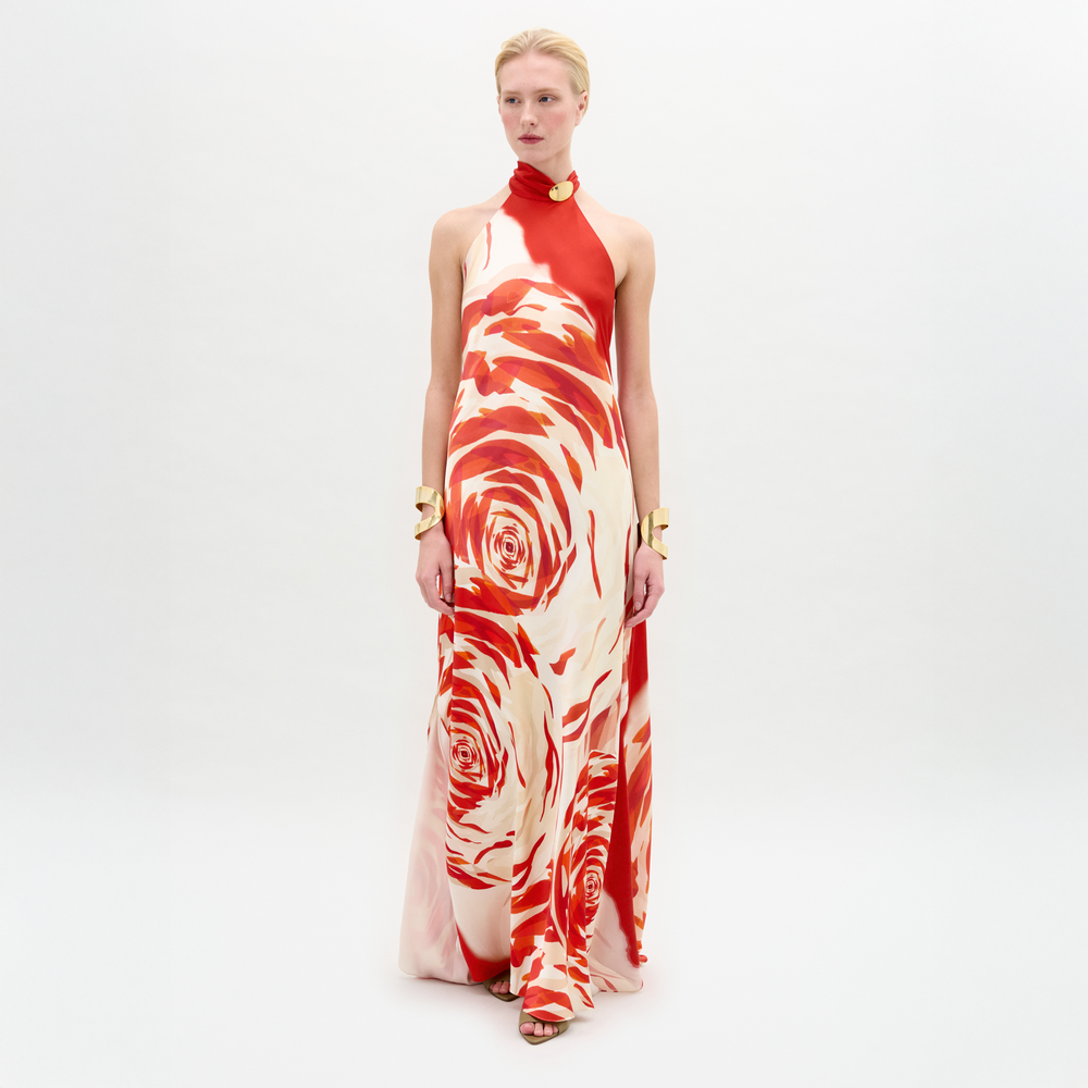 A person stands against a white background wearing the Sherry Dress—a red and white silk maxi with a bold swirling rose pattern, halter neckline, draped column fit—styled with gold accessories.