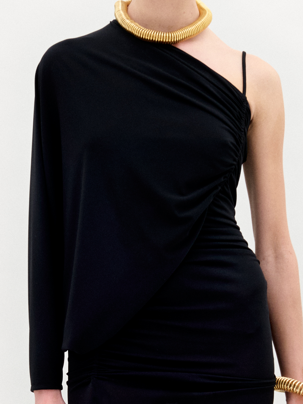 Against a plain background, a person showcases the Shiri Dress, a long black asymmetrical column silhouette crafted from Italian fabric, paired with gold accessories.