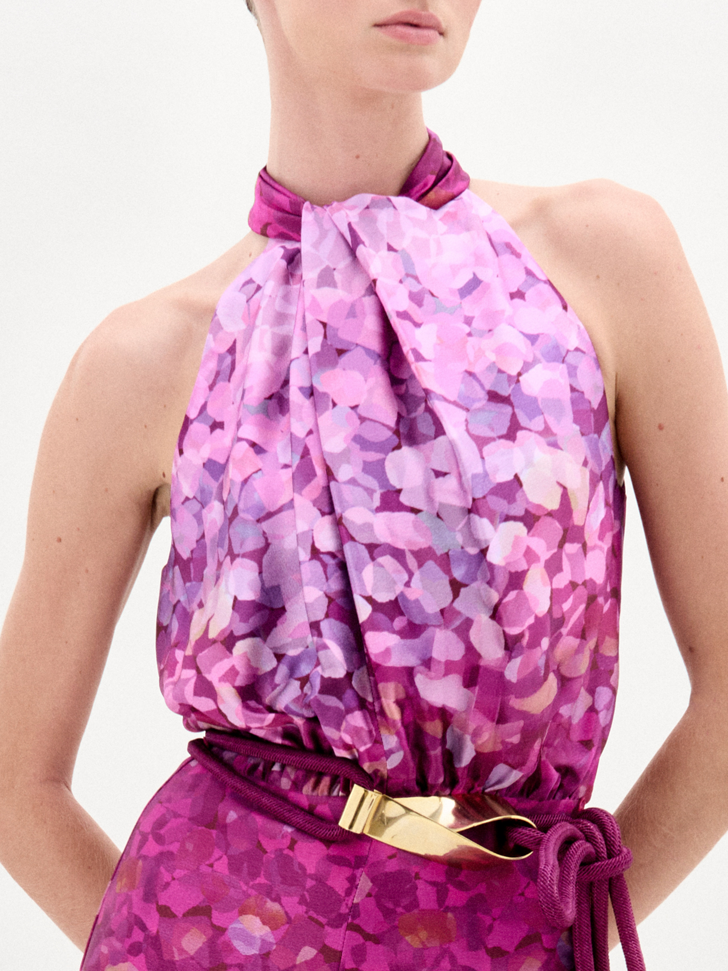 A woman stands against a plain white background wearing the Siv Jumpsuit, a sleeveless halter-neck style made from Italian fabric with a pink and purple abstract print and a matching belt.