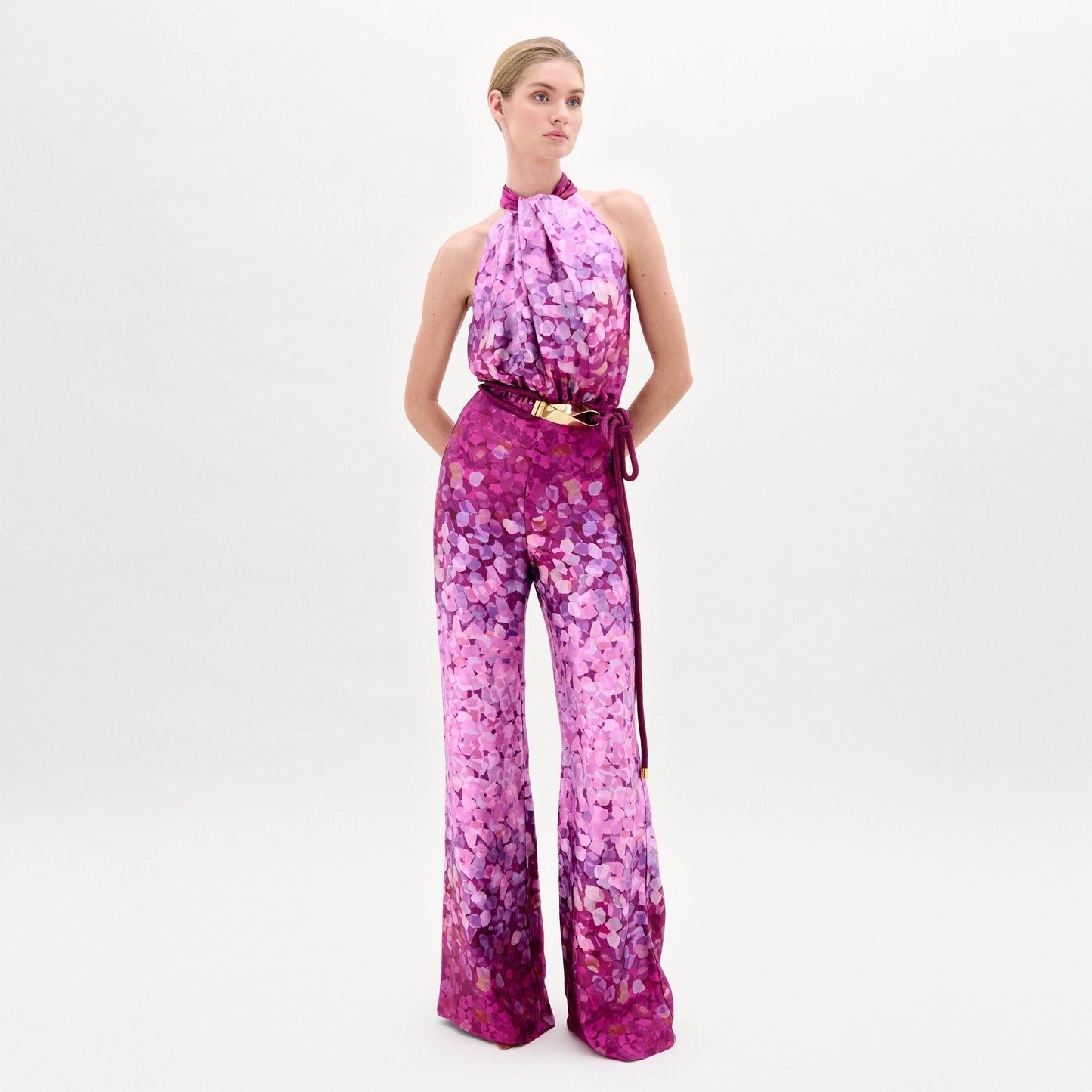 A woman stands against a plain white background wearing the Siv Jumpsuit, a sleeveless halter-neck style made from Italian fabric with a pink and purple abstract print and a matching belt.