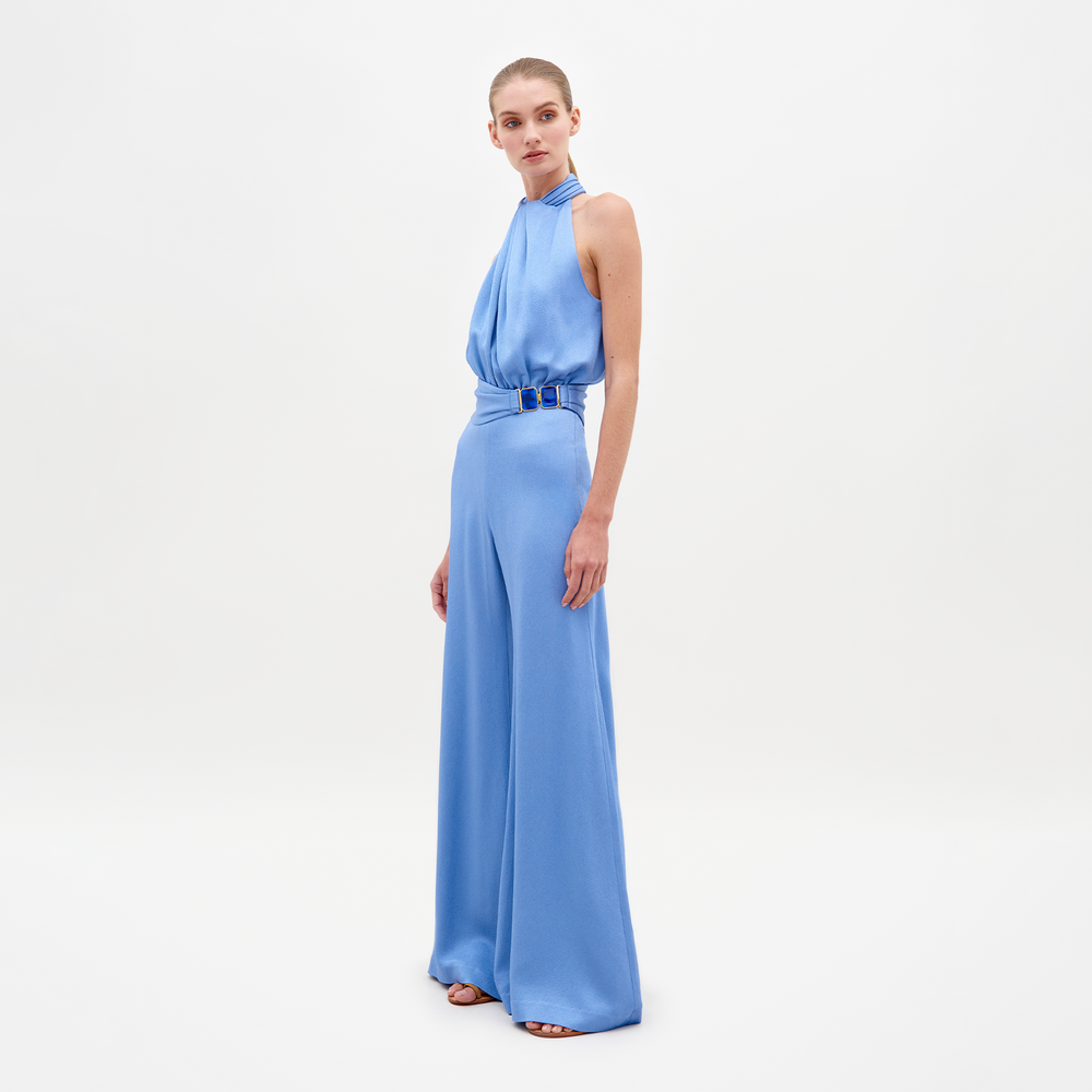 A person wearing the Siv Jumpsuit Periwinkle, featuring a sleeveless, light blue, halterneck design with wide-leg pants and a belted waist, stands against a plain white background. Pre-order now to ensure it ships by February 15th, 2025.