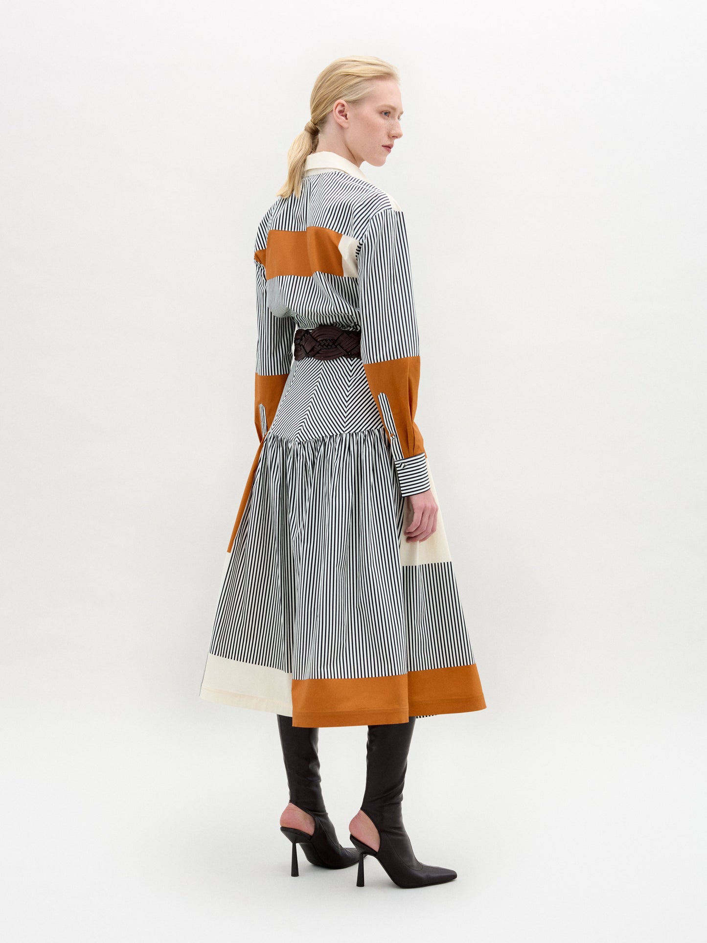 A woman with blonde hair and dark heeled boots stands slightly turned away, wearing the Shoreh Skirt—a gathered midi skirt in Italian fabric with vertical black-and-white stripes, orange-brown accents, and a dark belt—against a neutral background.