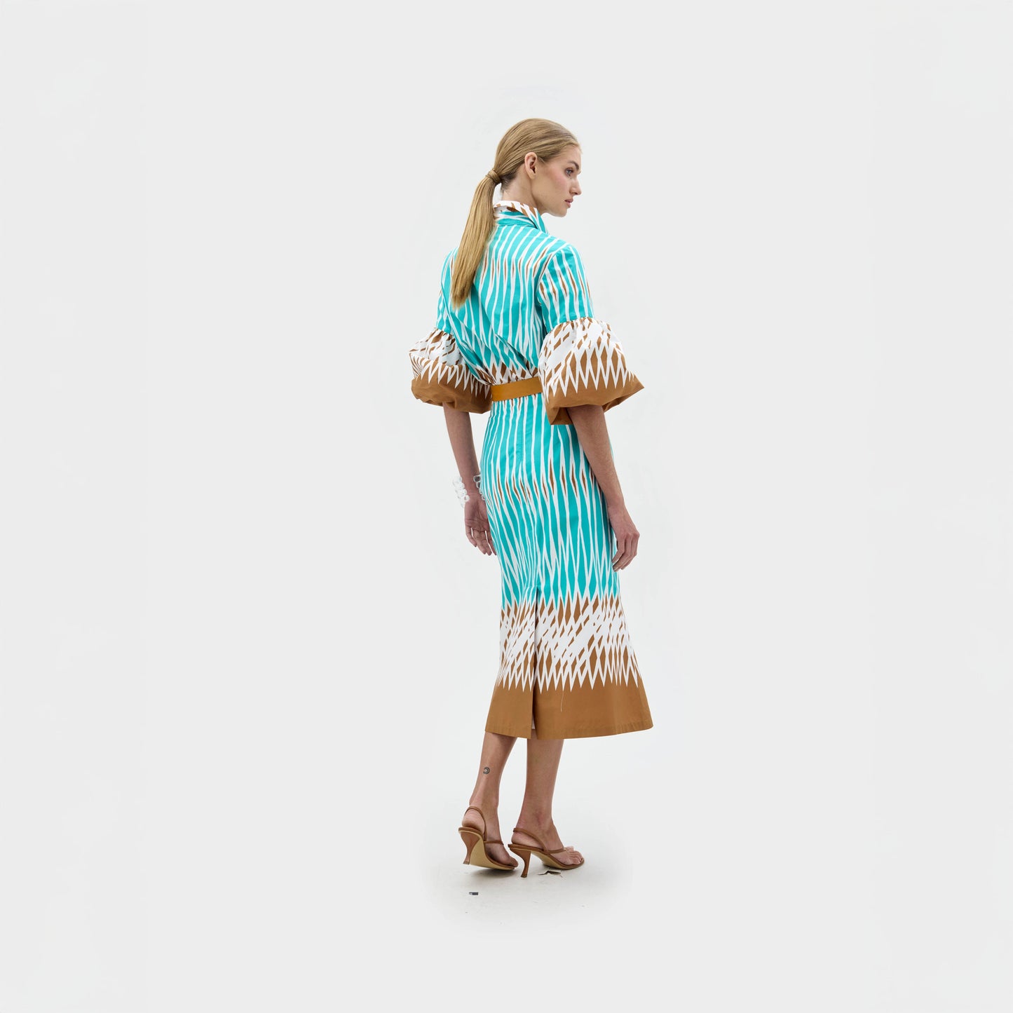 A woman stands against a plain white background wearing the Atira Skirt, a high-waisted, turquoise, white, and brown patterned midi skirt styled with puffed sleeves, a wide belt, strappy sandals, and bracelets.