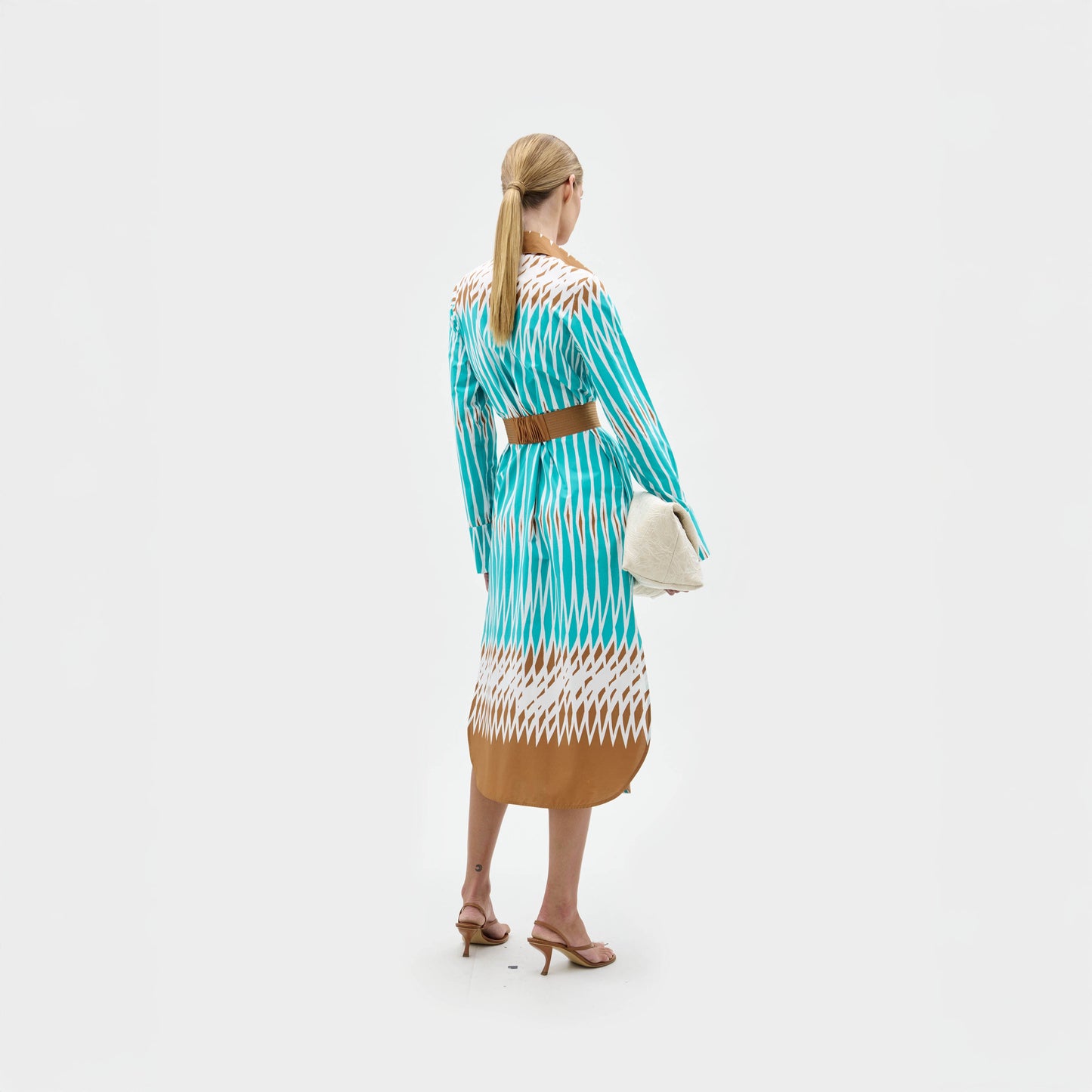 A woman stands against a plain background wearing the Ronzona Dress—a blue and brown patterned Italian midi dress—styled with a brown detachable belt, brown sandals, and holding a large white clutch.