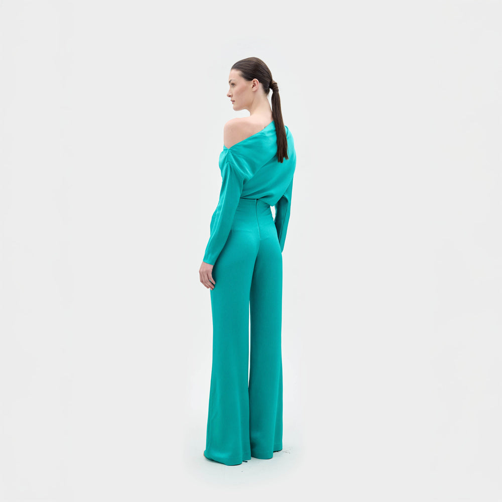 A woman stands against a plain background wearing the Palermo Pant, a teal Italian-fabric one-shoulder jumpsuit with wide-leg pants and a large tan belt.