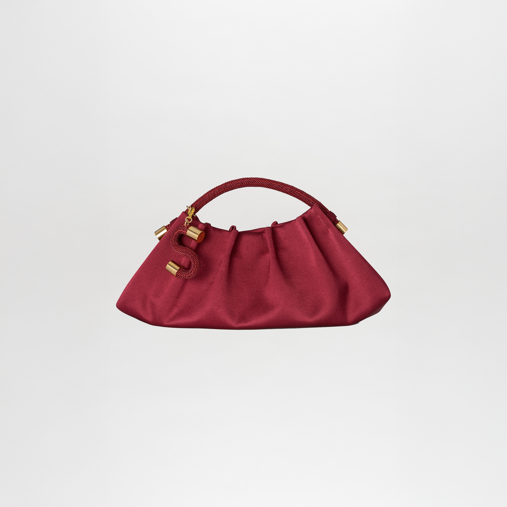 The Trapezius Clutch is a maroon pleated crepe handbag with gold accents, a textured handle, and a gold letter "S" charm.