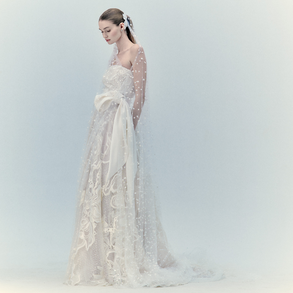A woman in the Selman Dress Pearl, a strapless Atelier maxi crafted from Italian textile with sheer lace and polka dot accents, stands against a pale background, looking down with her hands at her sides.