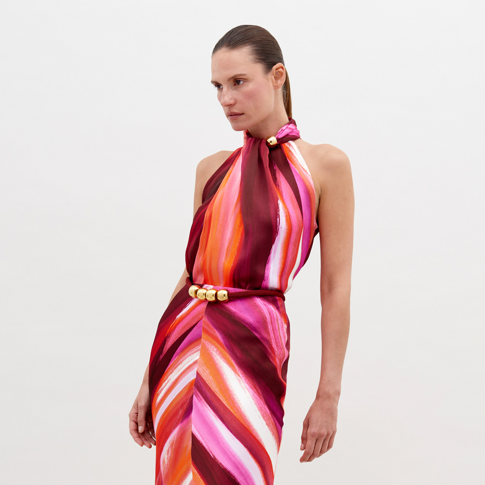 A woman stands against a plain background, wearing a halter-neck dress with a multicolored pattern of pink, orange, and red hues, accessorized with a gold belt buckle. Pre-order the Shela Blouse Magenta Abstract Waves now to ensure delivery by November 15th, 2024.