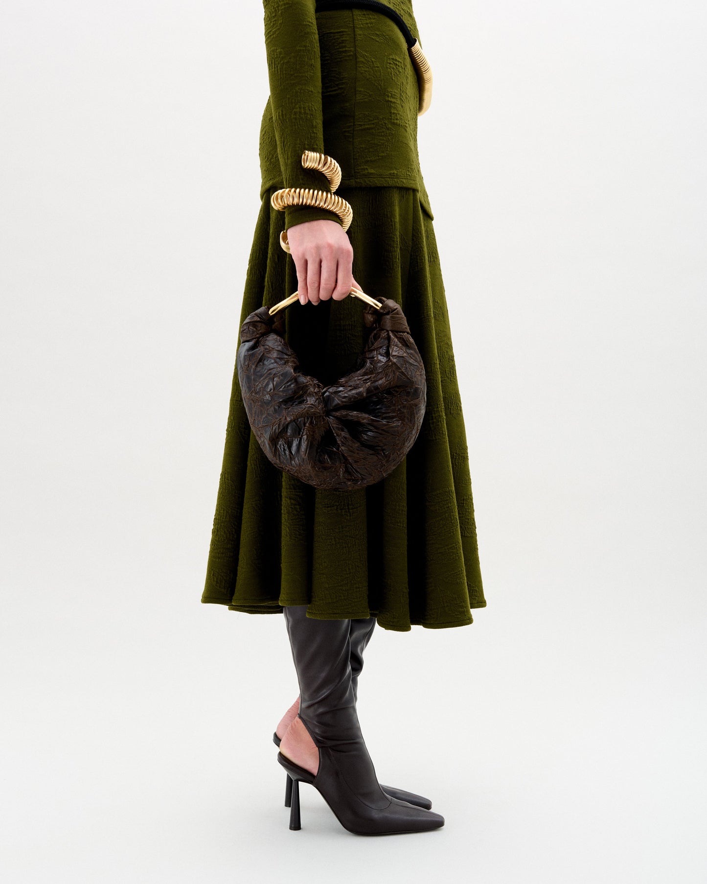 A person in an olive green textured dress, black heeled boots, gold spiral bracelets, and holding the Sofia Bag (Small)—a petite Italian fabric bag with a gold-toned handle—against a plain light background.