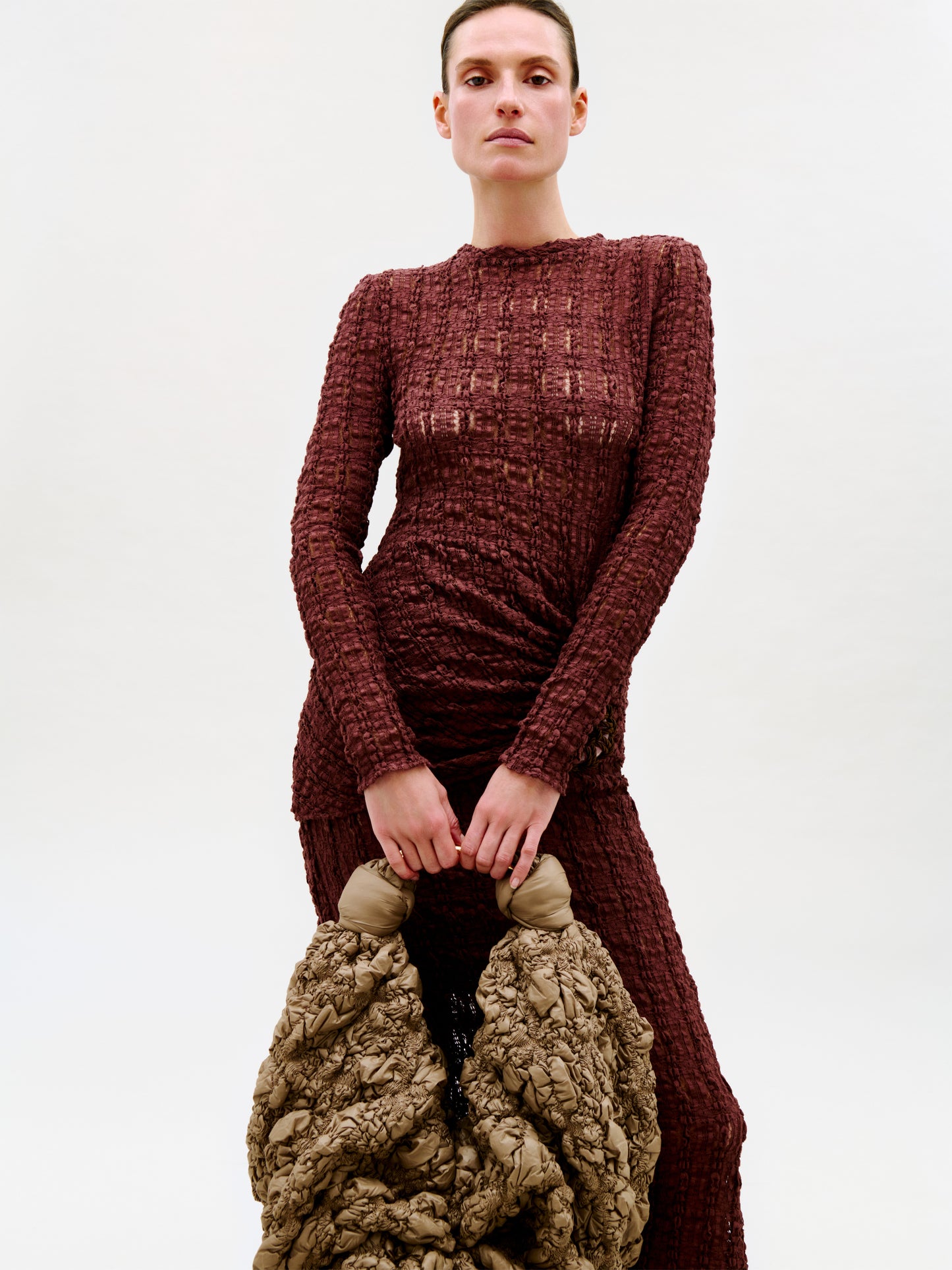 A person in a textured burgundy dress stands against a plain background, holding the Sofia Bag (Maxi), its gold-toned handle gleaming as they grip the Italian-crafted piece with both hands in front of them.
