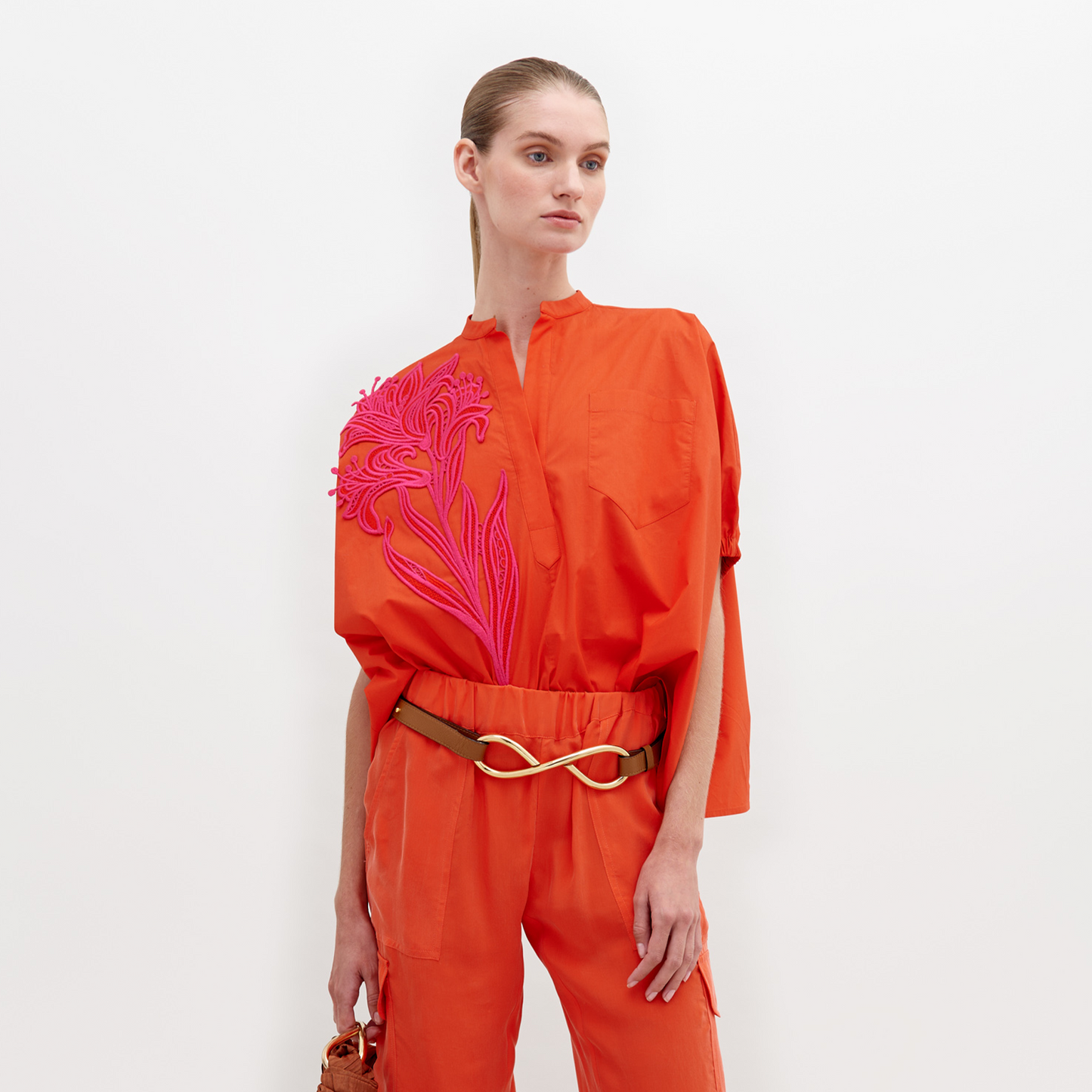 A woman stands against a white background, wearing an orange outfit with a pink floral pattern on the upper left side and a gold belt. The Susanne Blouse Scarlet is available for pre-order until November 15th, 2024.