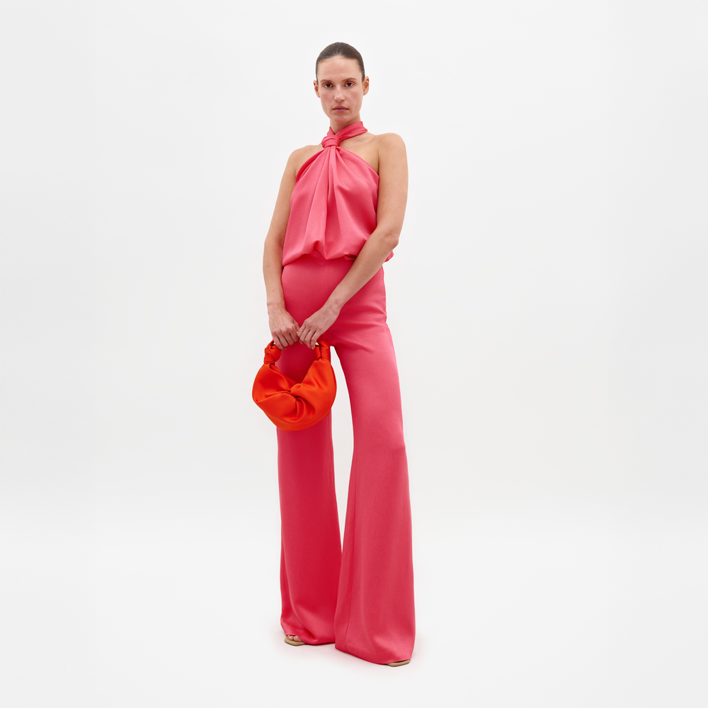 A person wearing a Palermo Pant Peony set, which includes a pink halter top and matching wide-leg pants, holds an orange handbag against a plain white background. This stylish ensemble is available for pre-order, with a ship by date of February 15th, 2025.