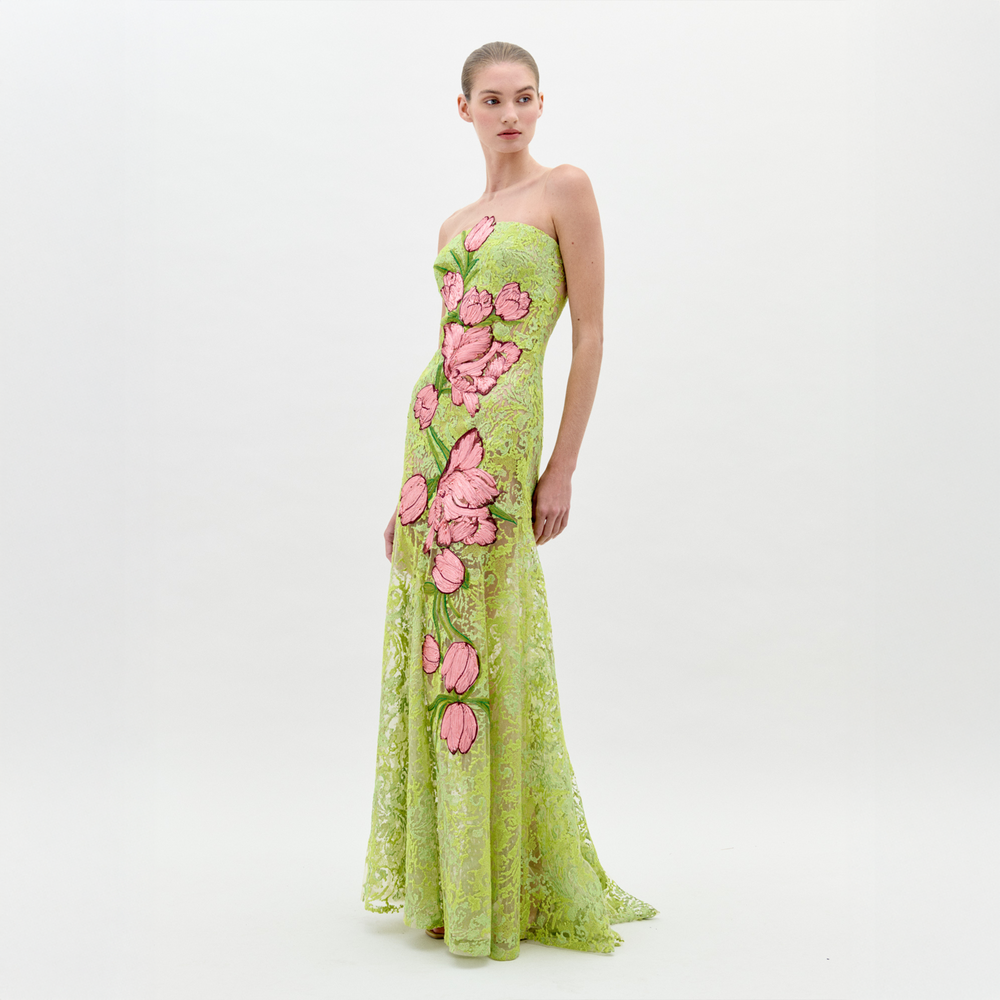 A woman stands wearing the Tulip Dress Rose Tulips, a strapless, green lace dress adorned with large pink floral designs, looking to her left against a plain light background. Pre-order now for exclusive access; shipping by February 1st, 2025.