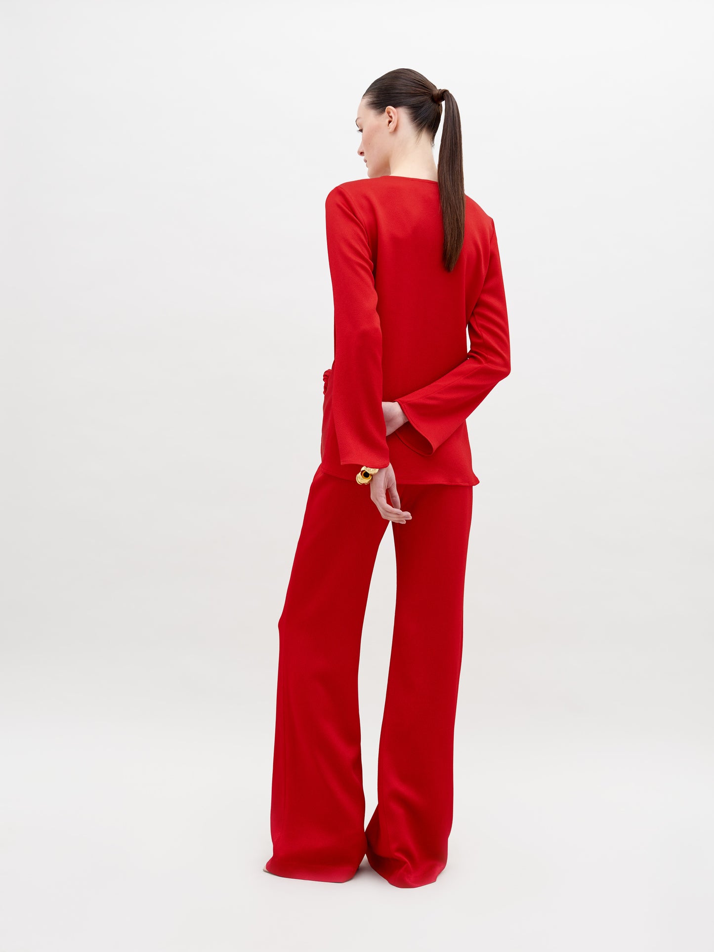 A woman with long dark hair in a ponytail stands with her back to the camera, wearing the Palermo Pant—a bright red high-waisted, wide-leg style crafted from luxurious Italian textile—set against a plain white background.