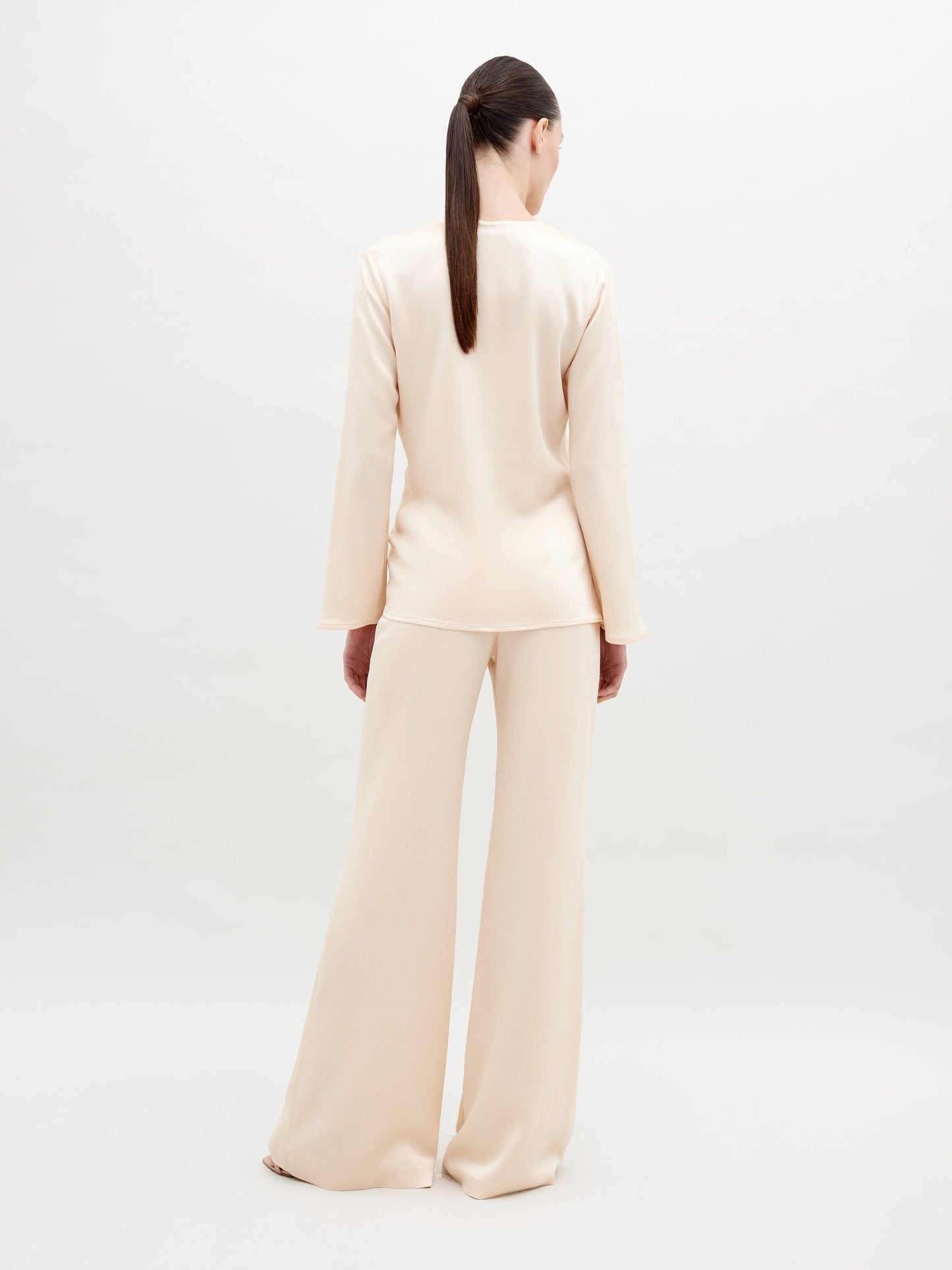 A woman with long brown hair in a low ponytail stands with her back to the camera, dressed in loose, cream-colored Palermo Pants made from luxurious Italian textile, featuring wide legs and long sleeves, against a plain white background.