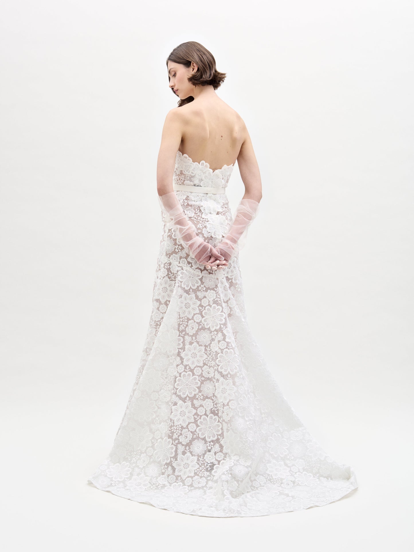 A woman poses against a plain white background in the Valencia Dress, which features a strapless design with lace floral patterns. She complements it with sheer gloves.