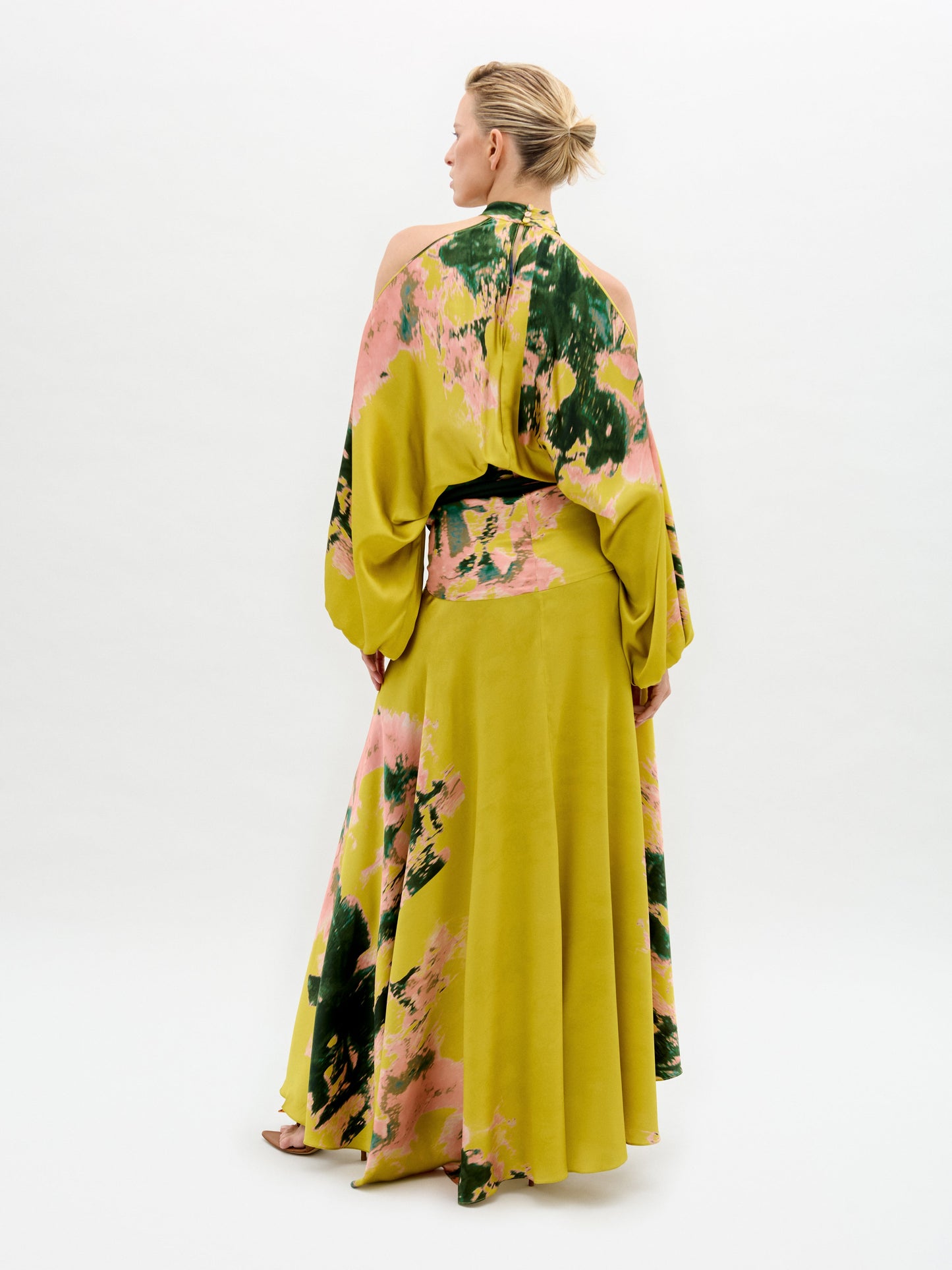 A person wears the Valeria Dress, a flowing maxi dress in yellow, pink, and green patterns with open shoulders and a 3D cord belt, standing against a plain white background.