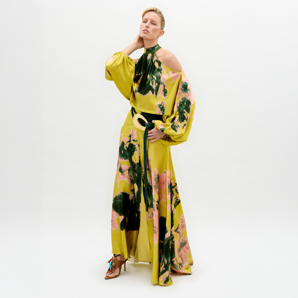 A person wears the Valeria Dress, a flowing maxi dress in yellow, pink, and green patterns with open shoulders and a 3D cord belt, standing against a plain white background.