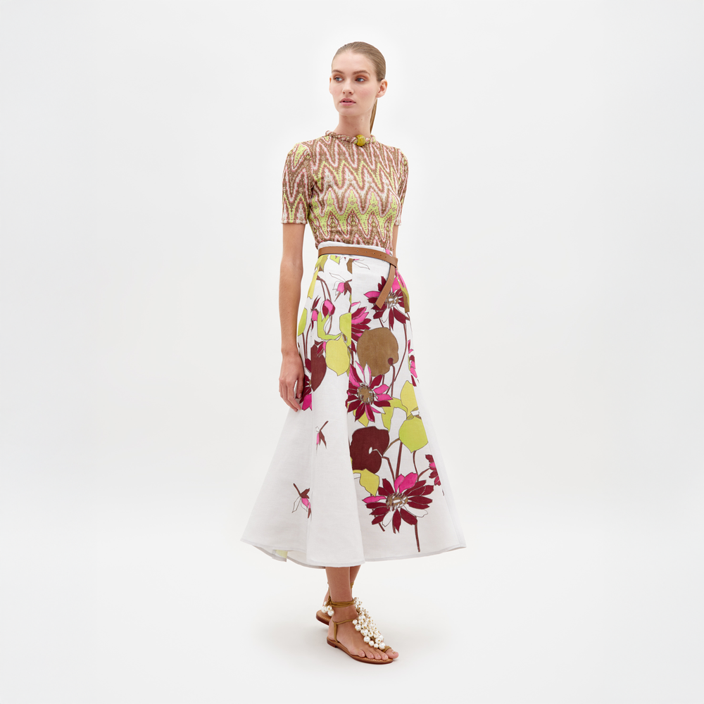 A person standing in a patterned top and the Erin Skirt Botanical Multifloral paired with sandal heels against a plain white background, ready for pre-order. Expected ship by date is February 15th, 2025.