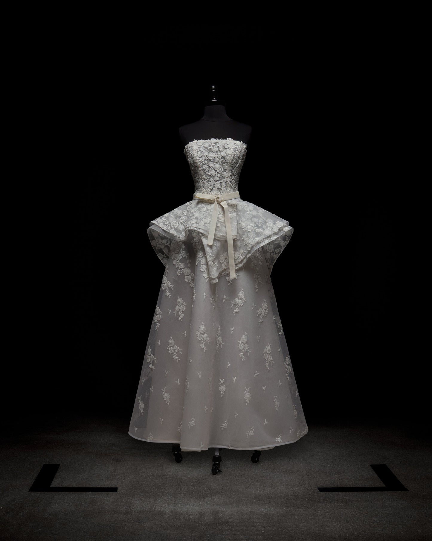 A woman in the Viviana Dress, a strapless white wedding gown featuring floral lace details and a peplum design, stands against a light plain background.