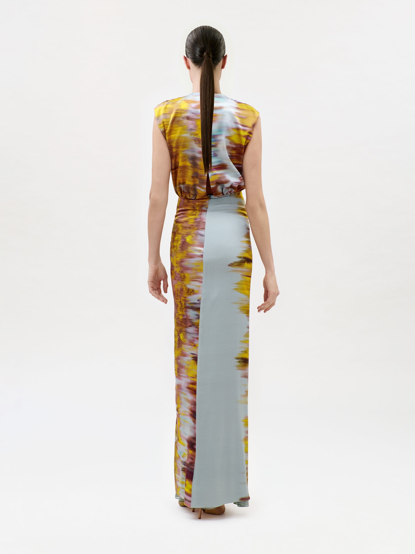 A woman with long brown hair in a ponytail models the Winona Dress, a sleeveless, floor-length gown featuring a yellow, brown, and pale blue tie-dye pattern. She is shown standing with her back to the camera against a white background.