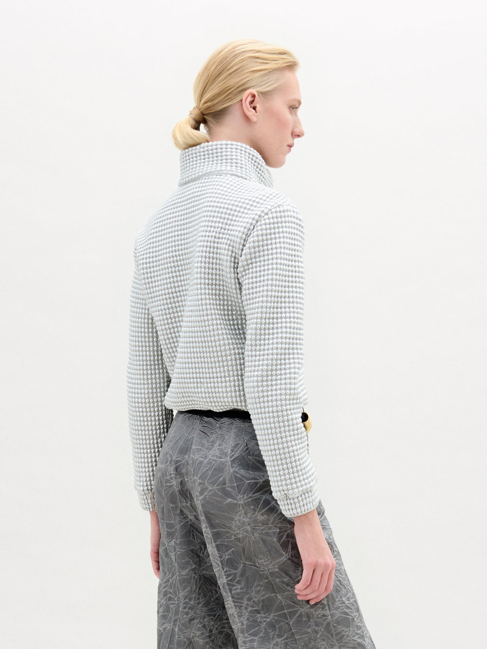 A person stands against a plain white background, wearing the oversized Zalta Jacket made from Italian fabric in a checkered pattern, paired with crinkled gray trousers and hands in pockets.