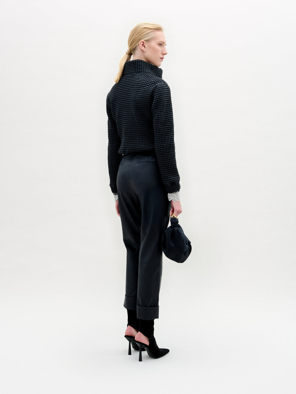 A person stands against a plain white background wearing the Zalta Jacket, a dark cropped piece crafted from Italian fabric, paired with a striped shirt, dark trousers with a gold belt, and holding a dark bag.
