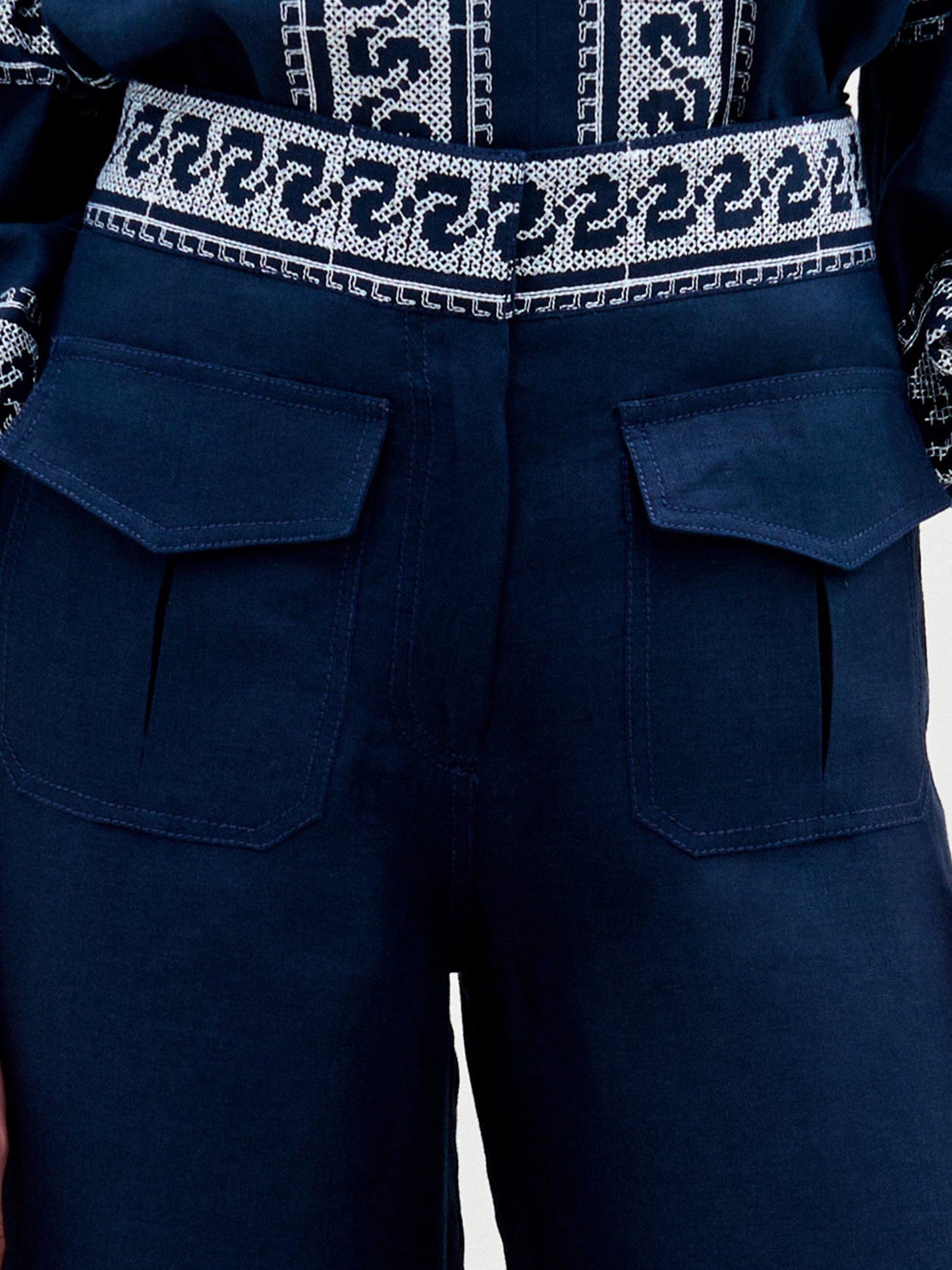 A woman stands against a plain background, wearing a navy blue embroidered blouse with ruffled sleeves and the Zina Pant—matching embroidered linen wide-leg pants—paired with gold sandals.