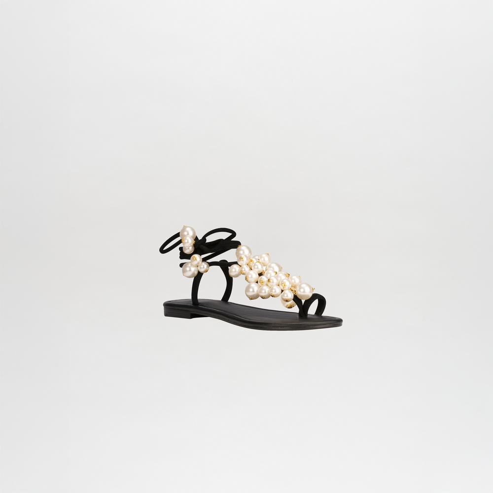The Zaira Sandals are chic black leather footwear featuring pearl-embellished straps, a flat sole, and elegant wrap-around ankle straps, all elegantly presented against a white background.