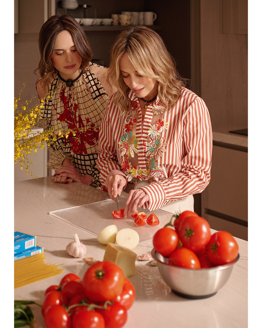 Two women prepare fresh ingredients in a warm kitchen: one slices tomatoes while the other watches, with bowls of produce nearby, creating a cozy, collaborative mood.