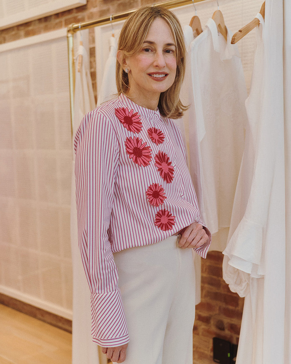 Silvia Tcherassi wearing a striped blouse with red floral embellishments and white pants stands in a chic boutique setting. She is smiling softly, exuding confidence