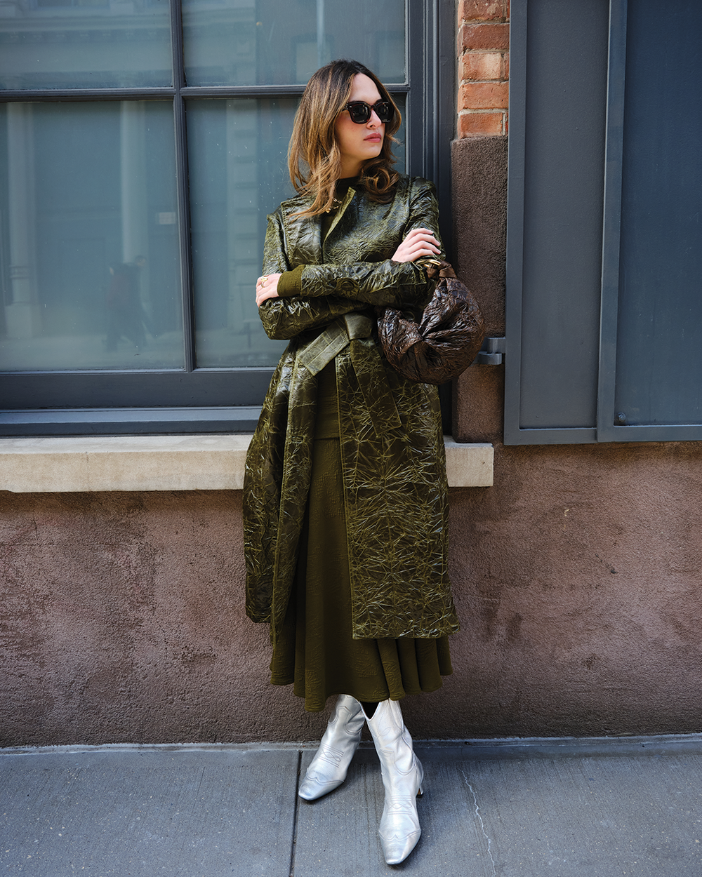 Sofia Tcherassi wearing a textured olive-green coat over a matching outfit stands against a building wall, arms crossed, wearing sunglasses and white ankle boots.