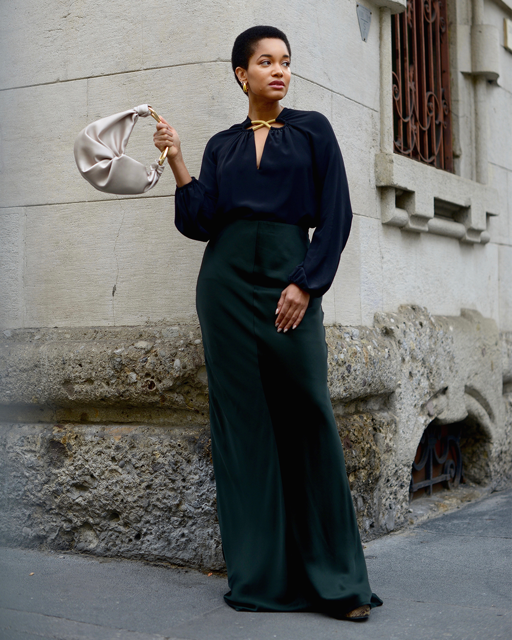 Tamu MchPherson stands confidently in a dark blouse and high-waisted wide-leg pants, holding a light handbag. The stone building background adds elegance.