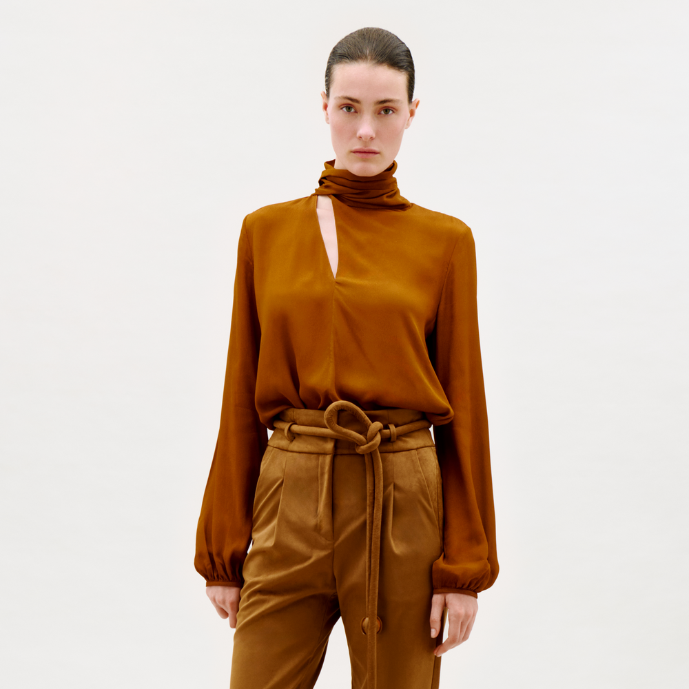 A person stands against a plain light background, wearing the Alanis Blouse—a copper brown long-sleeve top with a high neck and front slit—paired with matching high-waisted pants tied with a fabric belt.