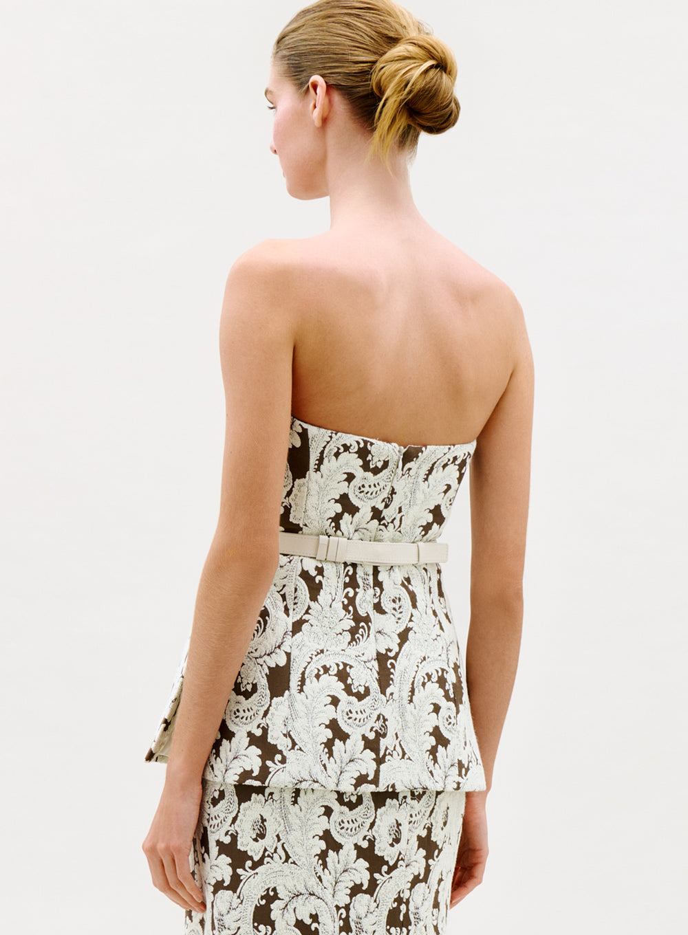 A woman with her hair in a neat bun is seen from the back, wearing the Astor Top, which features a strapless, fitted design with a white and brown ornate floral pattern and a thin white belt. The background is plain and light-colored.