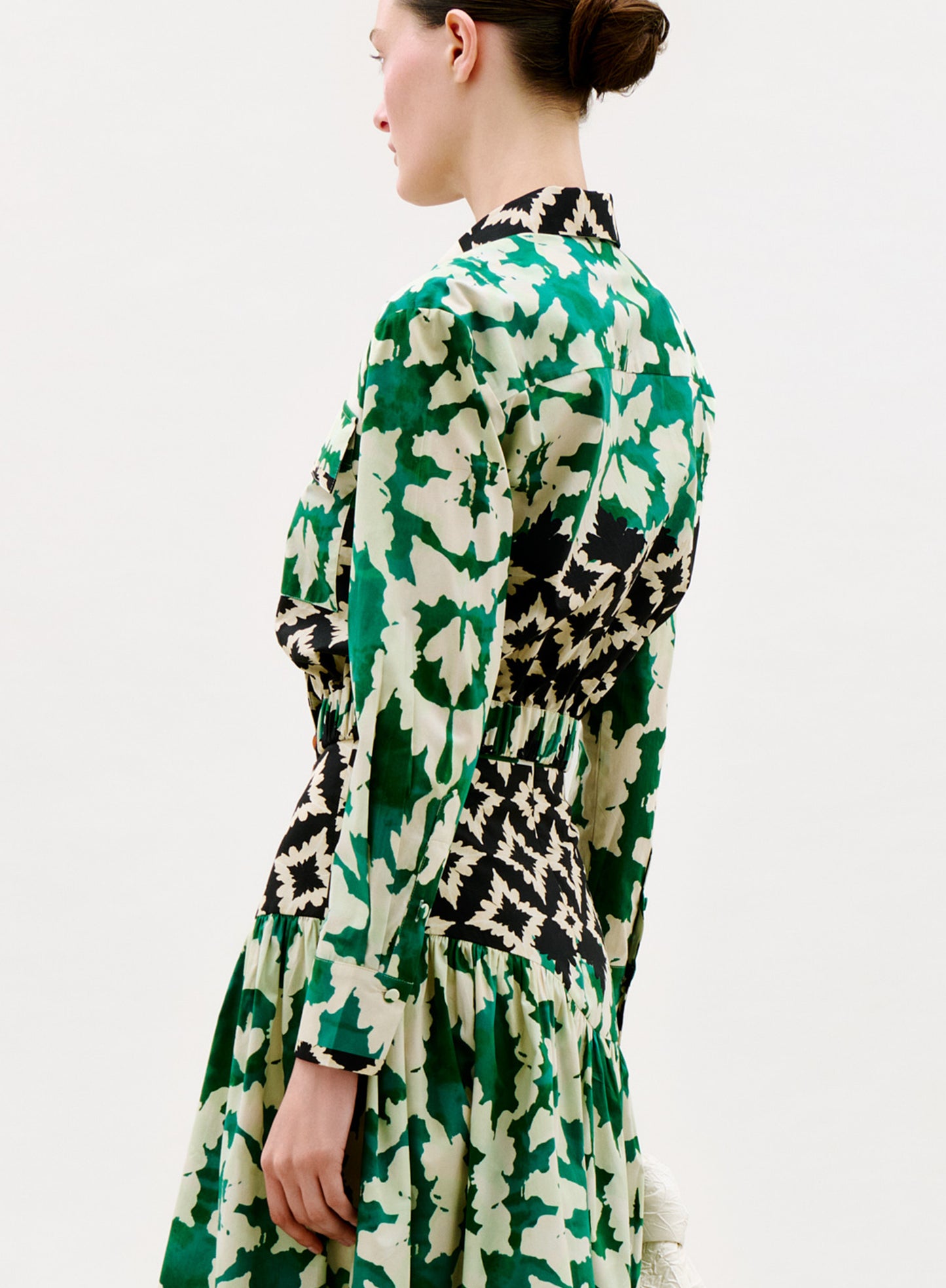 A woman stands sideways in the Soleil Blouse, which features a green and cream leafy pattern with black geometric accents. Her hair is in a neat bun, set against a plain white background.