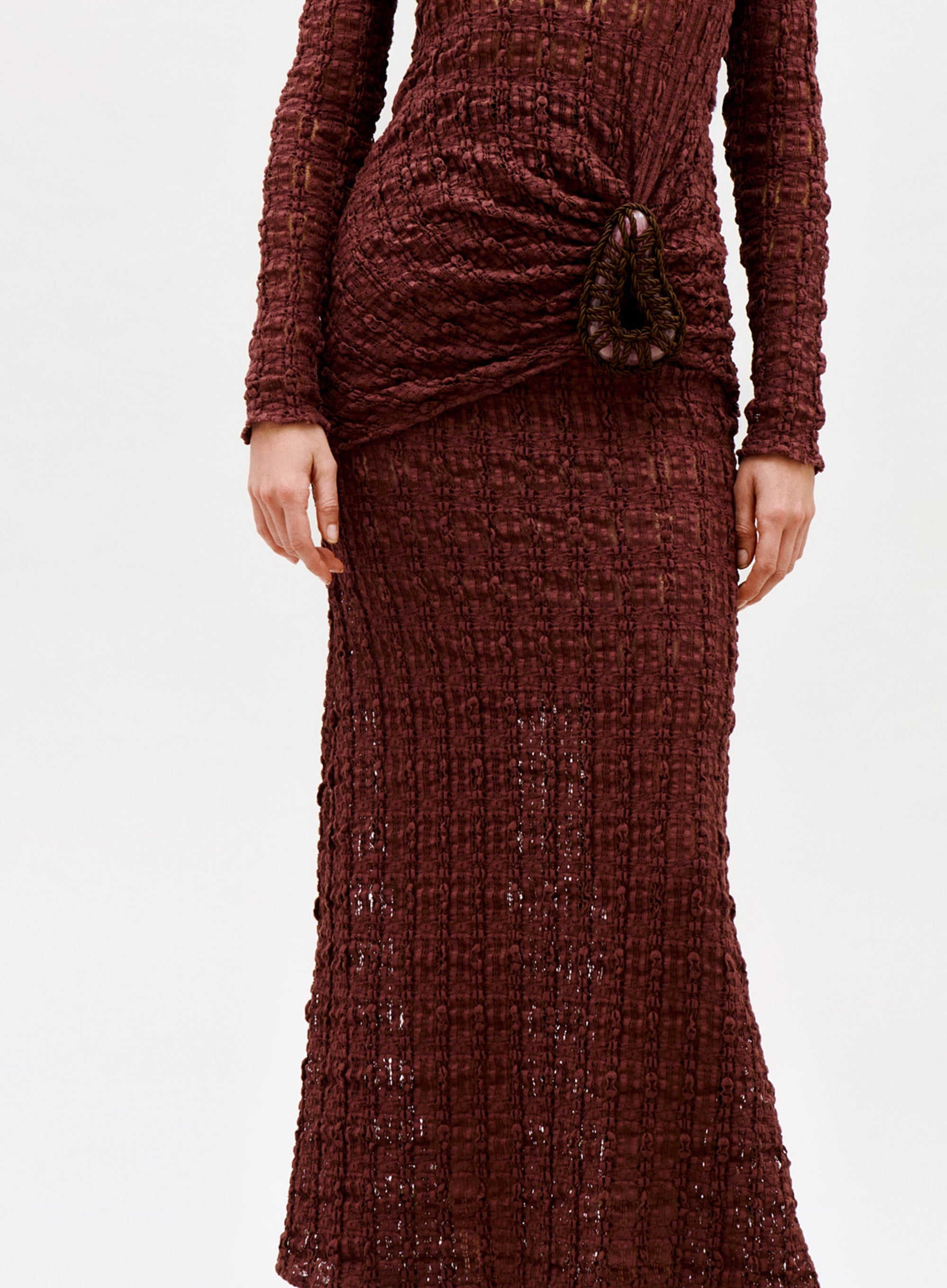 A person wearing the Ciji Skirt, a textured burgundy knit piece with a distinctive gathered hip detail and long sleeves. The photo shows the body from shoulders down against a plain light background.