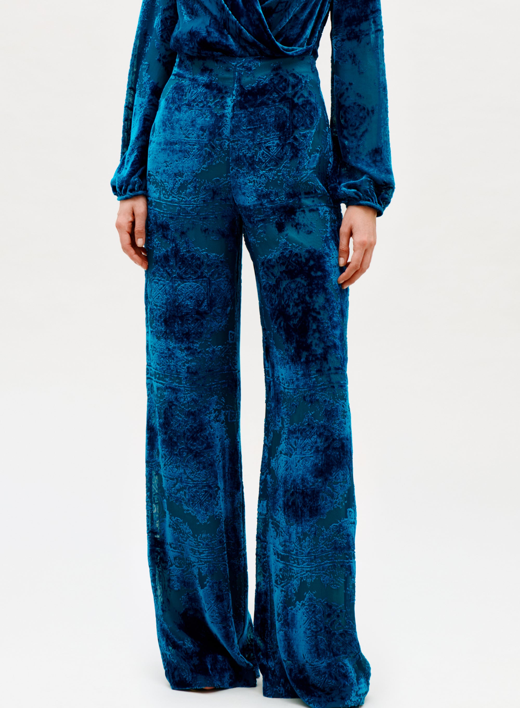 A person wears the Como Pant—high-waisted, wide-leg velvet pants in deep blue with a subtle textured pattern—paired with a matching long-sleeve top, set against a plain, light background.