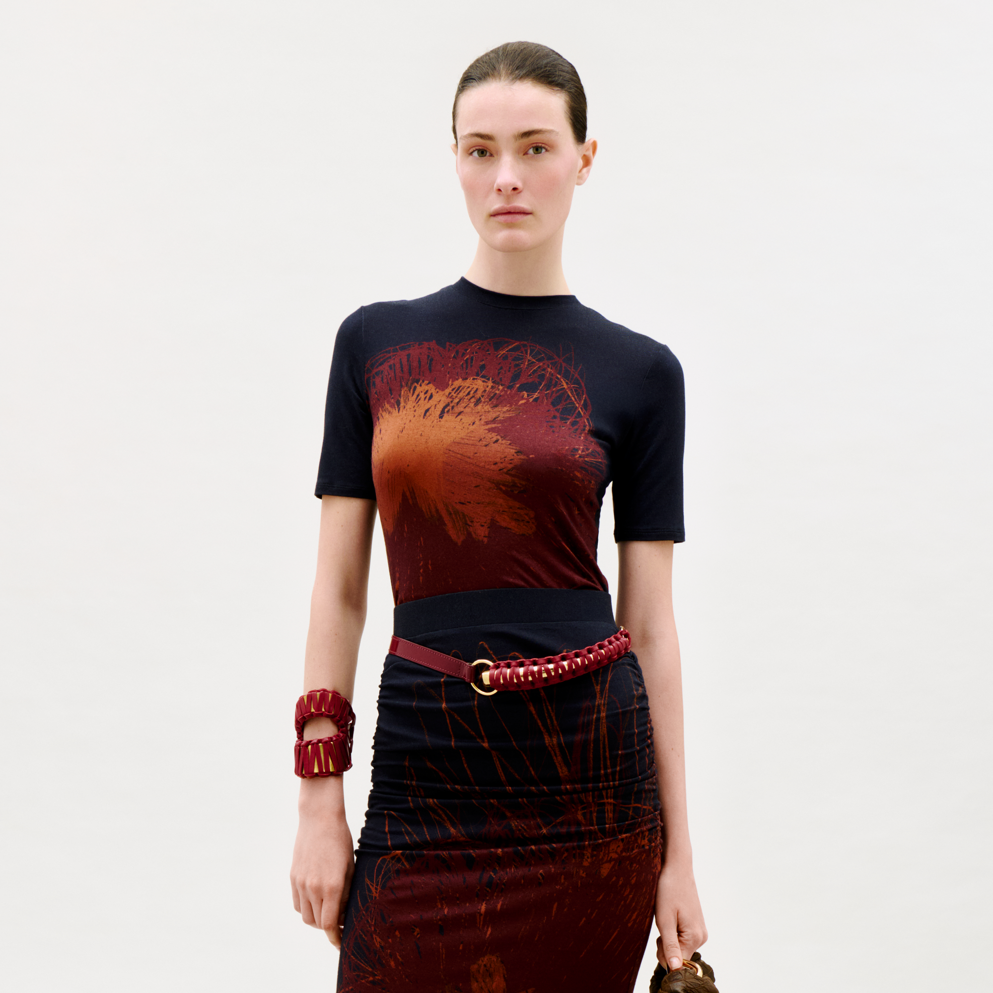 A woman stands against a plain light background, wearing the Fano T-Shirt, paired with a red braided belt and chunky red bracelet, holding a handbag and looking directly at the camera.