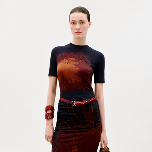 A woman stands against a plain light background, wearing the Fano T-Shirt, paired with a red braided belt and chunky red bracelet, holding a handbag and looking directly at the camera.