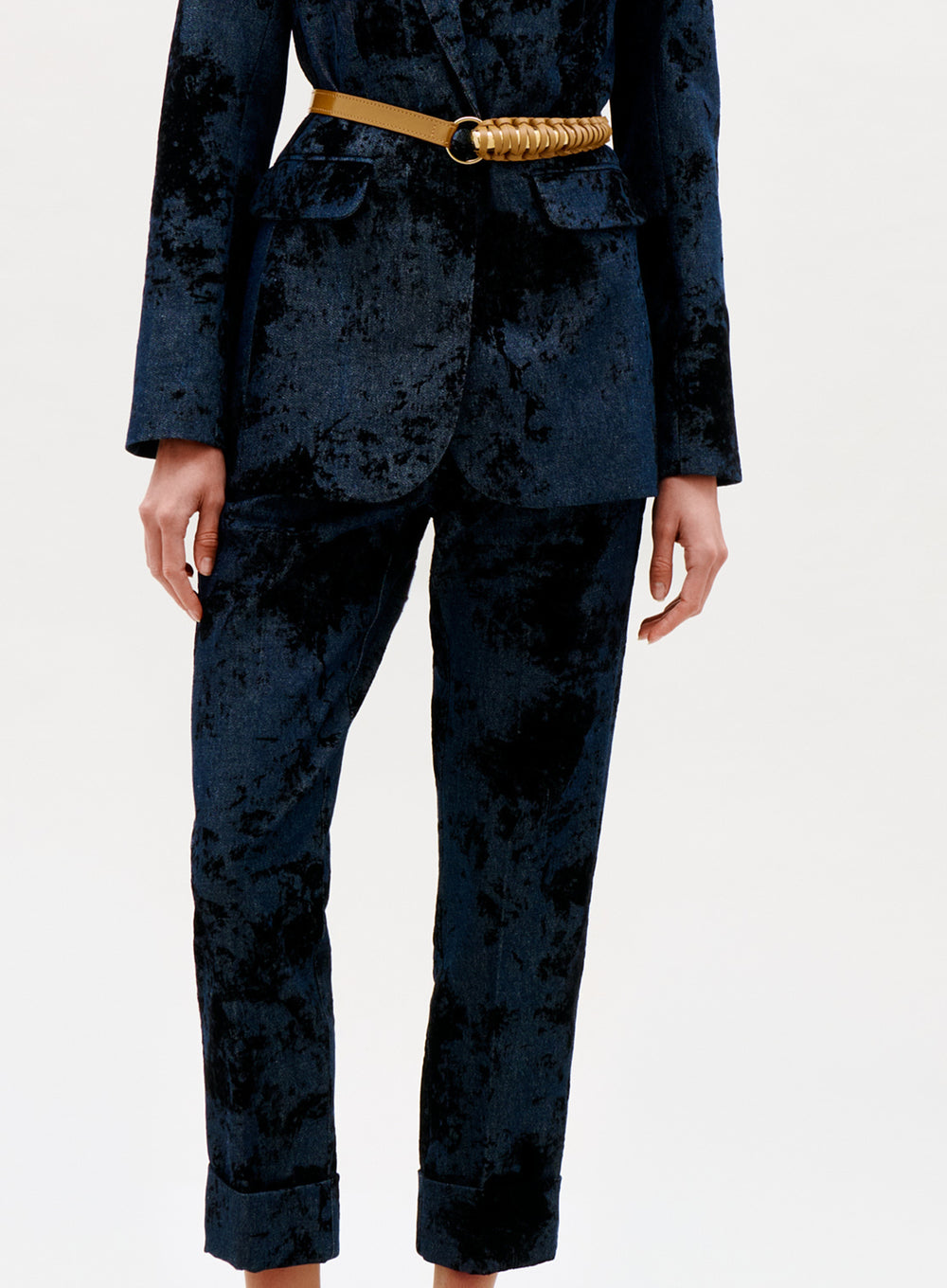 A person showcases the Moad Pant in a dark blue velvet suit with a gold chain belt, standing against a plain light background. Only the body from the shoulders down is visible; the suit features a soft, textured look.