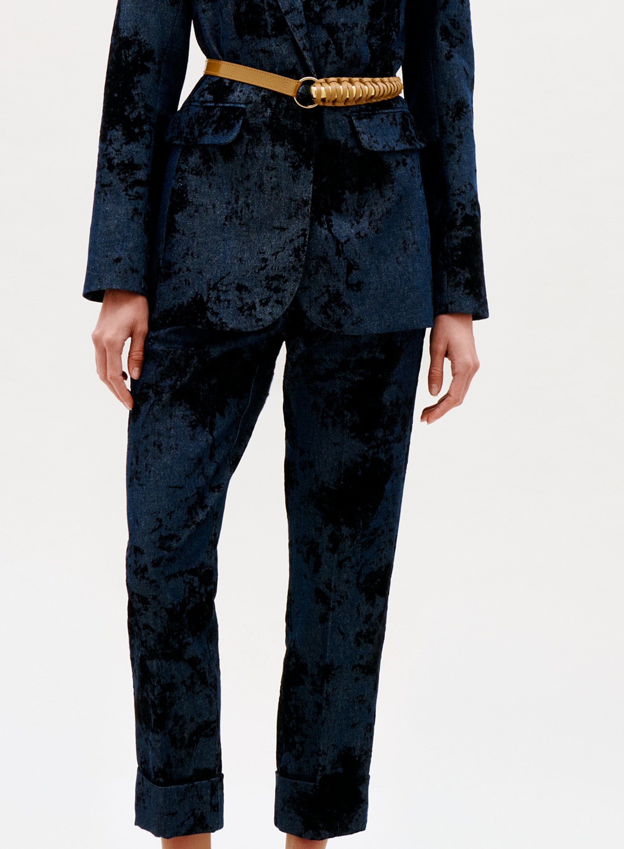 A person showcases the Moad Pant in a dark blue velvet suit with a gold chain belt, standing against a plain light background. Only the body from the shoulders down is visible; the suit features a soft, textured look.