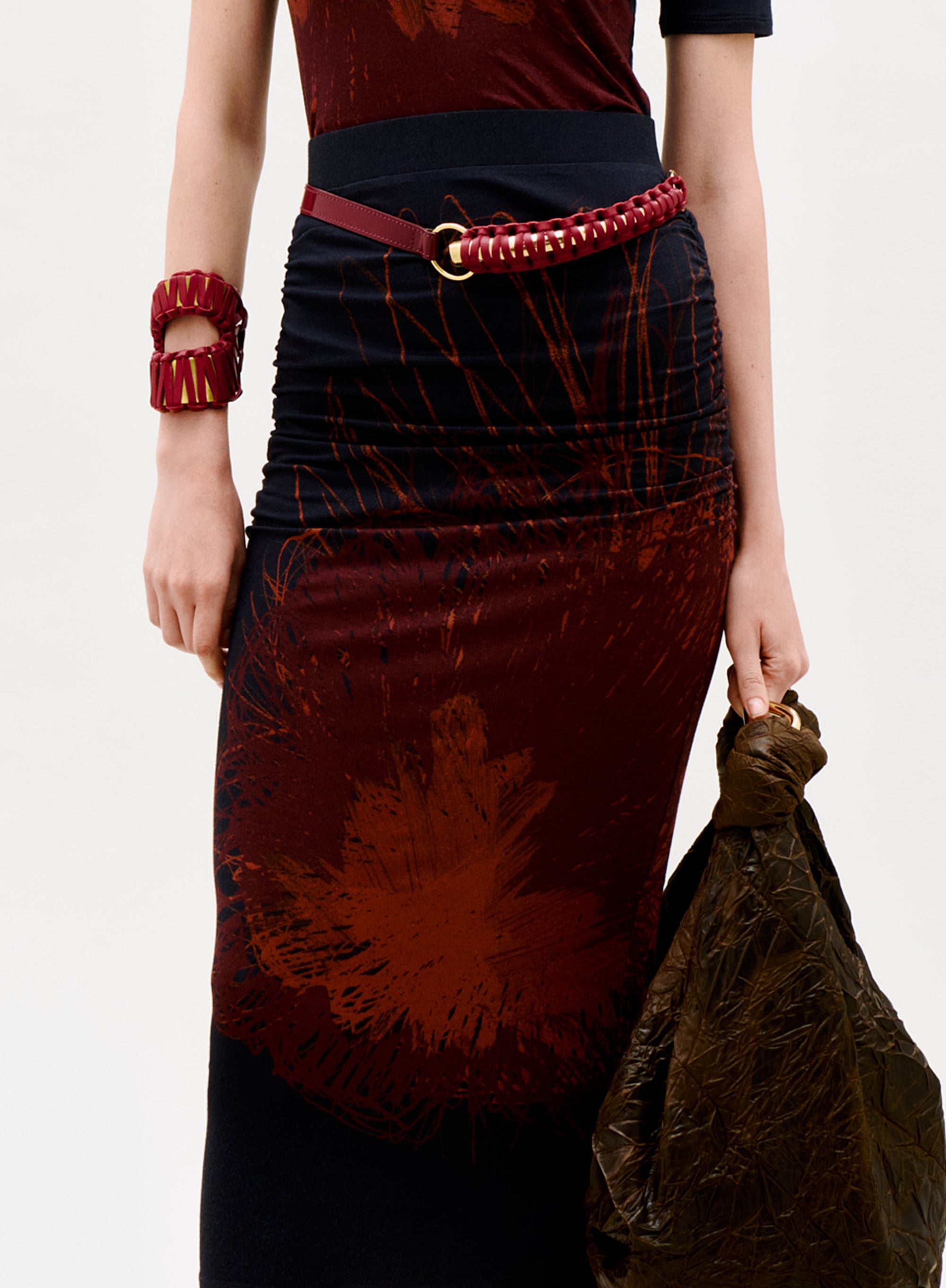 A person wears the Niove Skirt with red and dark abstract patterns, styled with a red braided belt, matching bracelet, and holding a crinkled dark brown bag against a plain light background.